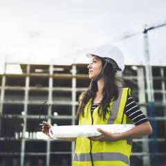 Junge Frau mit Helm und Warnweste auf Baustelle, hält Baupläne. Sie verfolgt ihren Weg im Bauingenieurwesen berufsbegleitend.