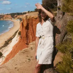 Kobieta w lnianej szmizjerce z paskiem, oparta o drzewo na klifie z widokiem na plażę. Idealne szmizjerka stylizacje na wakacje.