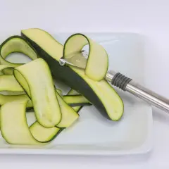 Powstaje makaron z cukinii. Spiralne paski zielonych warzyw i obieraczka na białym talerzu.