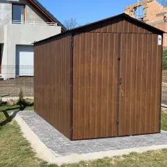 Nowy domek narzędziowy w brązowym kolorze, gotowy na budowę. Ustawiony na kostce brukowej, obok domu w budowie.
