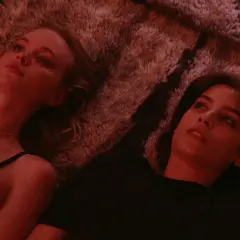 Två unga kvinnor ligger nära varandra på en fluffig matta, en i svart linne, den andra i svart t-shirt. En scen från en threesome serie.