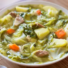 Bol de soupe au chou vert, pommes de terre, carottes et viande, avec du pain à côté.