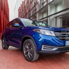Niebieski SUV DFSK Seres 3 na tle nowoczesnej architektury.