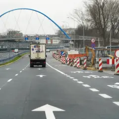 Droga w budowie, z koparką i barierami. W tle niebieski most i **aleja** z ruchem samochodowym.