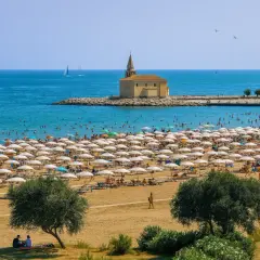 Vue aérienne d'une plage bondée à Caorle, avec des rangées de parasols blancs, des baigneurs dans la mer bleue et une petite église sur un îlot rocheux.