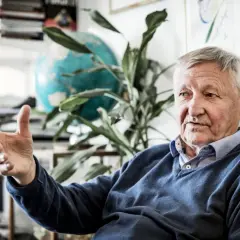 Äldre man med grått hår, liknar Ragnar Skanåker, gestikulerar med handen. Bakom honom syns en jordglob och växter.
