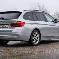 Srebrne BMW Seria 3 kombi z polskimi tablicami rejestracyjnymi stoi na mokrej nawierzchni, otoczone suchą trawą i nagimi drzewami.