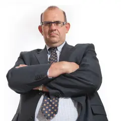 Mężczyzna w garniturze i okularach ze skrzyżowanymi ramionami, patrzący na wprost.