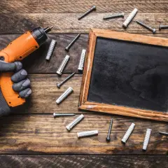 Un guante gris sujeta un taladro naranja, rodeado de varios tipos de herramientas como tornillos y tacos, junto a una pizarra sobre madera.