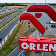 Duży znak ORLEN nad stacją paliw przy autostradzie. W tle parking z ciężarówkami i samochodami.