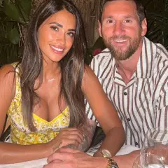 Uśmiechnięta Antonela Roccuzzo, żona Messiego, w żółtej sukience, siedzi obok niego przy stole.