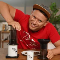 Mann mit rotem T-Shirt zeigt die Aeropress Zubereitung mit zwei Geräten und Kaffeebohnen.