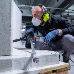 Operaio con mascherina, occhiali e cuffie usa un martello perforatore per fare un foro da 100 nel muro di cemento.
