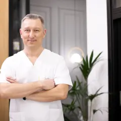 Uśmiechnięty lekarz w białej koszuli z rękami skrzyżowanymi na piersi, stoi w nowoczesnym wnętrzu.