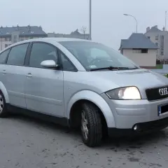 Srebrne Audi A2 z włączonymi światłami, zaparkowane na tle nowoczesnych budynków mieszkalnych.