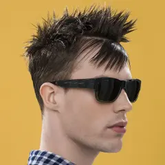Uomo con capelli a spazzola corti, occhiali da sole scuri e camicia a quadri blu e bianchi su sfondo giallo.