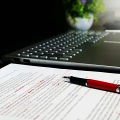 Czerwony długopis leży na dokumencie z poprawkami, obok laptopa. Widać tu różne odmiany cudzysłowu, które autor tekstu musiał poprawić.