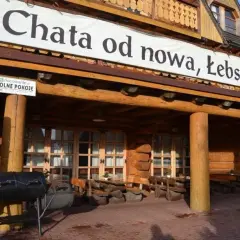Drewniana, łebska chata z banerem "od nowa, Łebskiej Chaty". Przed wejściem grill i stoliki.