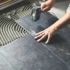 Mano che posa una piastrella scura su cemento fresco, con un martello per un posizionamento perfetto e rettificato.
