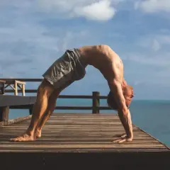 Mężczyzna wykonuje pozycję urdhva dhanurasana na drewnianym tarasie z widokiem na ocean i niebo.