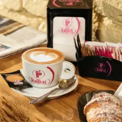 Un cappuccino senza schiuma, con un cuore di latte art, accompagnato da un croissant e un giornale.