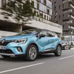 Un coche azul phev Renault Captur circula por una calle urbana moderna, con edificios de cristal y hormigón al fondo.