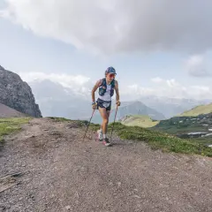 Corredor/a de montaña en un sendero rocoso, con bastones y vistas alpinas. Esto es trail running, pura aventura.