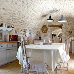 Cuisine rustique au style shabby chic, avec murs en pierre, table dressée et chaises anciennes.