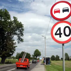 Znak zakazu wyprzedzania i ograniczenie prędkości do 40 km/h. Pomarańczowy pick-up z drabiną jedzie po drodze.