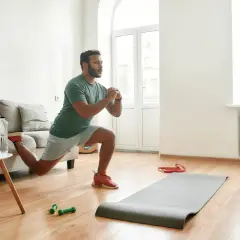 Un homme fait des fentes dans son salon, une idée pour comment commencer le sport à la maison. Un tapis, des haltères et un ordinateur sont à proximité.