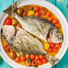 Orata all'acqua pazza, due pesci interi cotti in un brodo saporito con pomodorini e erbe aromatiche, serviti in un piatto bianco su un tavolo di legno blu.