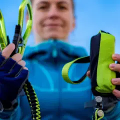 Ręce w rękawiczkach trzymają zielone karabinki i woreczek na via ferrata sprzęt.