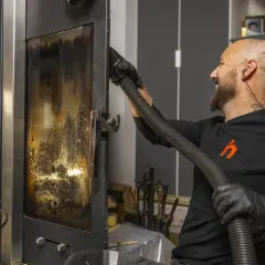 Un homme nettoie la vitre d'un poêle avec un aspirateur. Un bon entretien garantit la bonne fréquence de ramonage.