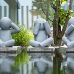 Drei graue Skulpturen von sitzenden Figuren, die ihre Ohren, Augen und Mund bedecken, spiegeln sich in einem Teich.