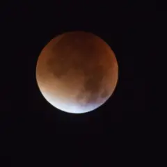 Czerwona poświata na Księżycu podczas zaćmienia. Widoczny fragment tarczy Księżyca w odcieniach pomarańczu i brązu na tle czarnego nieba.