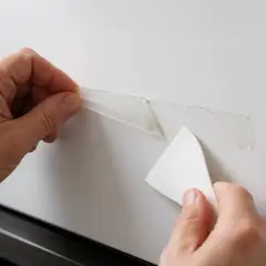 Dłonie usuwają taśmę dwustronną z białych mebli za pomocą plastikowej skrobaczki.