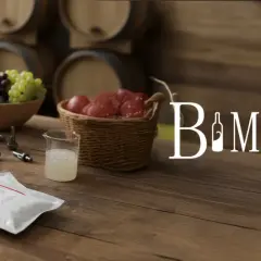 Na stole leżą opakowania z pożywką dla drożdży winiarskich, obok butelki wina, winogrona i beczki.