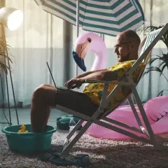 Mężczyzna w hawajskiej koszuli pracuje na laptopie, siedząc w leżaku z nogami w misce. W tle dmuchany flaming i parasol plażowy. Czas wykorzystać zaległy urlop!