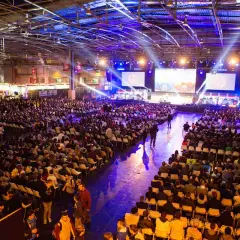 Gran multitud en un evento de convencion, sentada en sillas frente a pantallas gigantes con luces de escenario.