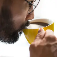 Man med glasögon och skägg dricker ur en gul mugg. Kanske känner han yrsel av kaffe.