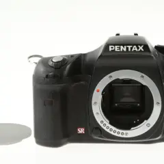Czarny aparat Pentax K10 z widocznym mocowaniem obiektywu i symbolem SR. Obok leży biała zaślepka. Ten aparat posiada zaawansowany ccd.