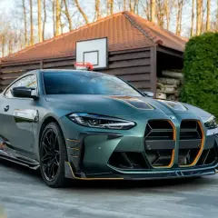Matowy, zielony BMW M4 G82 z pomarańczowymi akcentami, zaparkowany przed drewnianym domkiem z boiskiem do koszykówki.