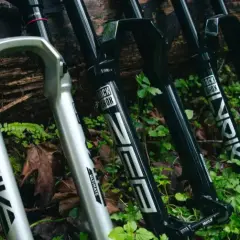A silver RockShox Pike and black RockShox Lyrik forks lean against a log, ready for trail adventures.