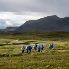 Hikers with blue backpacks trek through a vast, green landscape in Sweden, reminiscent of the Fjällräven Classic. Tents dot the distant hills under a dramatic sky.