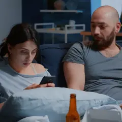 Para siedzi na kanapie, kobieta patrzy w telefon, mężczyzna obok niej wygląda na zamyślonego.