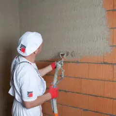 Tynkarz w białym ubraniu nakłada tynk maszynowy na ścianę z pustaków. Dowiedz się, ile kosztuje metr kwadratowy tynku maszynowego.