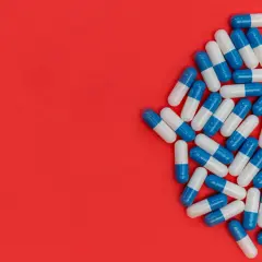 Kapsułki antybiotyku Amotaks w kolorze niebiesko-białym rozsypane na czerwonym tle.