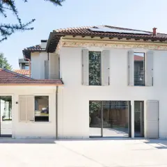 Casa con facciata color sabbia tortora, tetto in tegole rosse e pannelli solari. Ampie vetrate e persiane chiare.