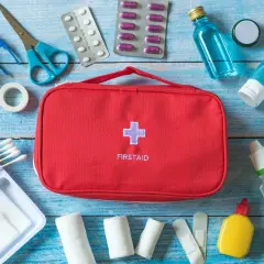 Czerwona apteczka z krzyżem i napisem FIRSTAID otoczona lekami, bandażami i akcesoriami medycznymi.