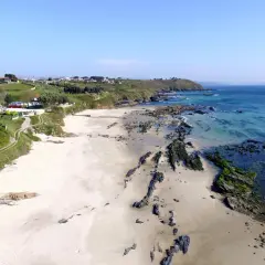 Vista aérea de la playa pragueira, con arena blanca, rocas cubiertas de algas y el mar azul turquesa. Casas y colinas verdes al fondo.